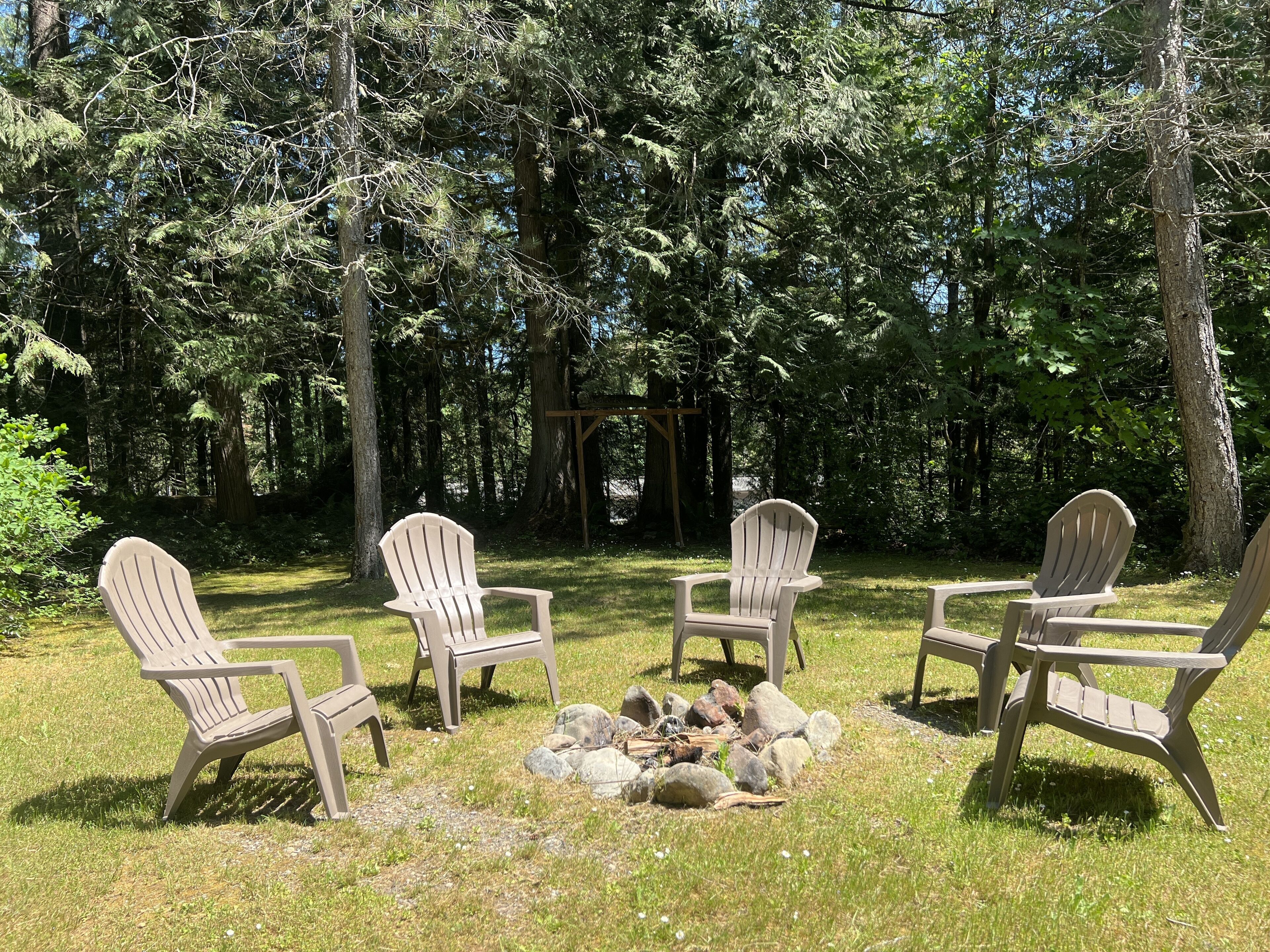A group of Adirondack chairs arranged around a small fire pit in a forested area.