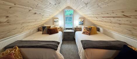 A cozy attic bedroom with two beds, a window, and a wooden ceiling.