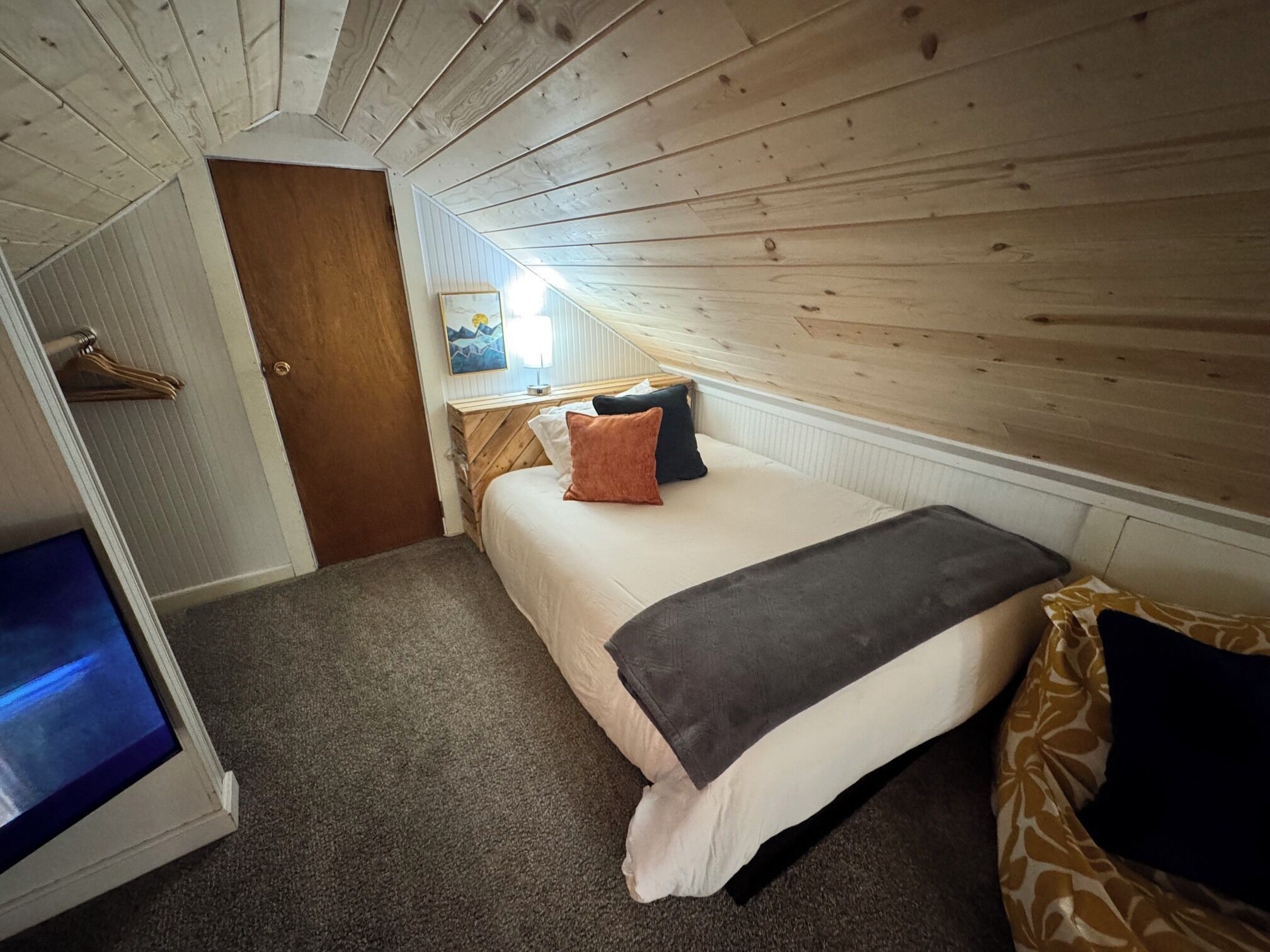 Un dormitorio con cama, mesita de noche, lámpara, techo de madera, puerta y televisor.