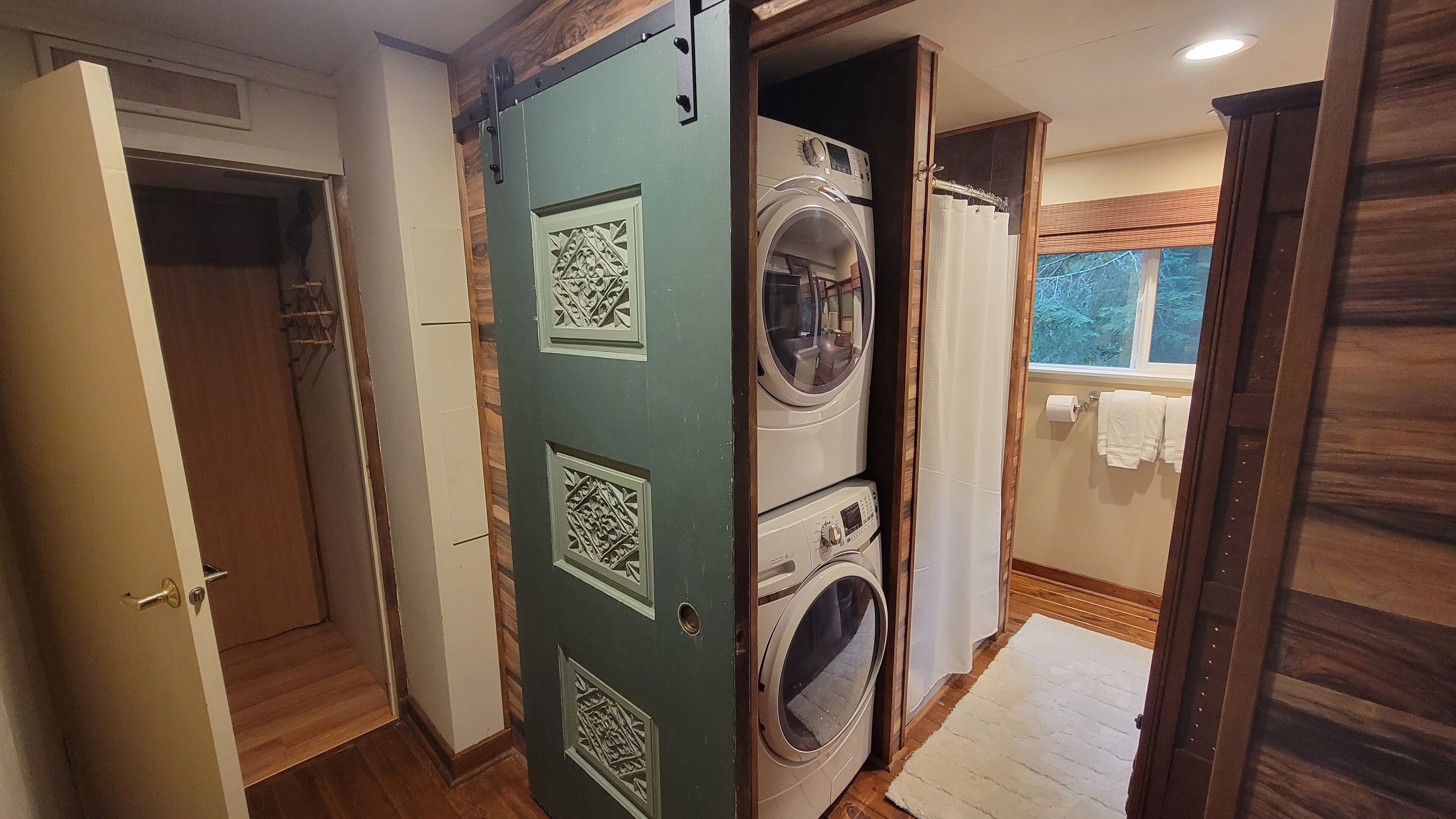 A laundry room with a stacked washer and dryer, a wooden door with decorative panels, and a window with a view of trees.