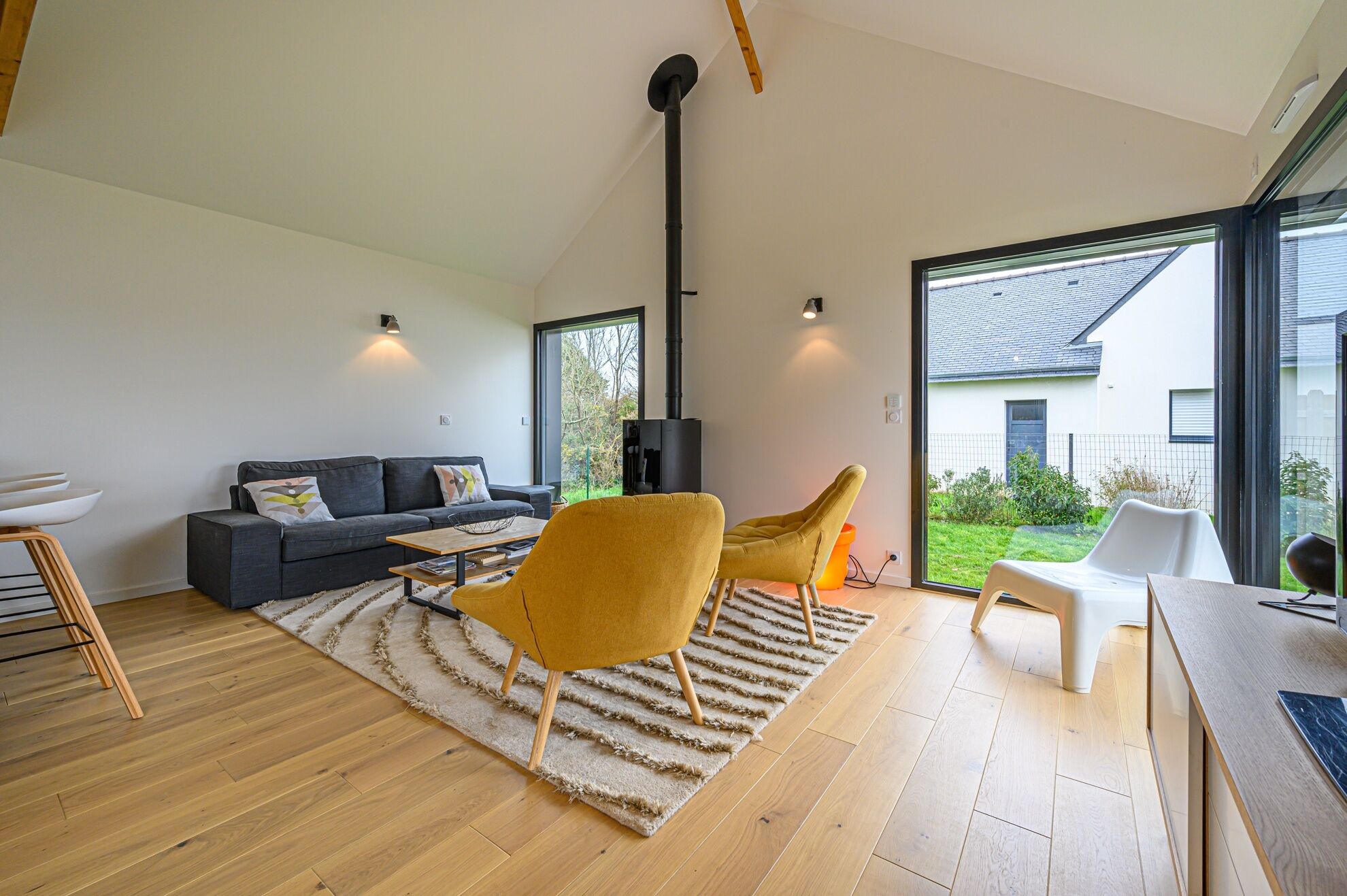 A modern living room with a sofa, two chairs, a coffee table, a rug, and a large window with a view of a house and garden.