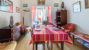 Een eethoek met een lange tafel, stoelen en een rood gestreepte tafelkleed. Er is een houten kast, een lamp en een raam met gordijnen.