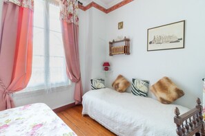 Une chambre à coucher avec un lit, une tête de lit en bois, une fenêtre avec des rideaux roses, une étagère murale agrémentée d’objets décoratifs et un tableau encadré.