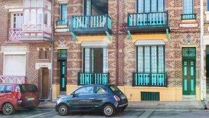 Een meerhoog gebouw met een bakstenen gevel, meerdere balkons met zwarte leuningen, en een kleine auto ervoor geparkeerd.