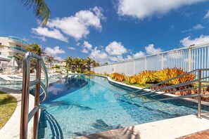 A clear blue swimming pool with a curved edge, surrounded by palm trees and tropical plants. - Apart na Praia de Cotovelo por Carpediem (Parnamirim)