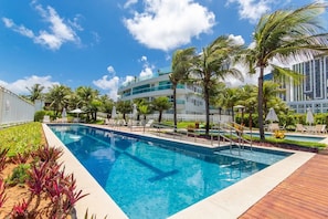 A swimming pool with a wooden deck, palm trees, and a modern building in the background.