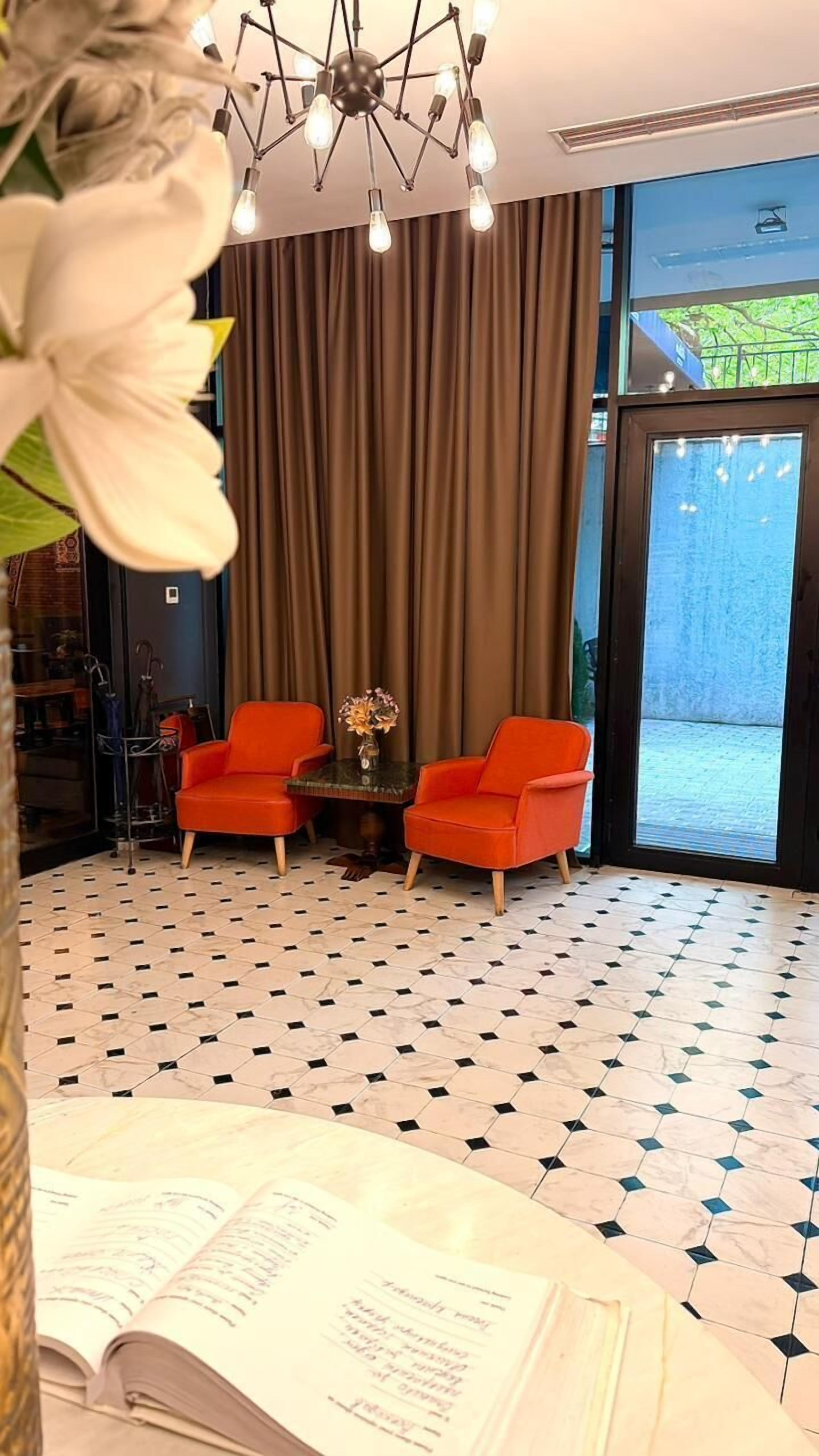Una habitación con dos sillas naranjas, una mesita con un florero, un suelo estampado y un ventanal con cortinas.