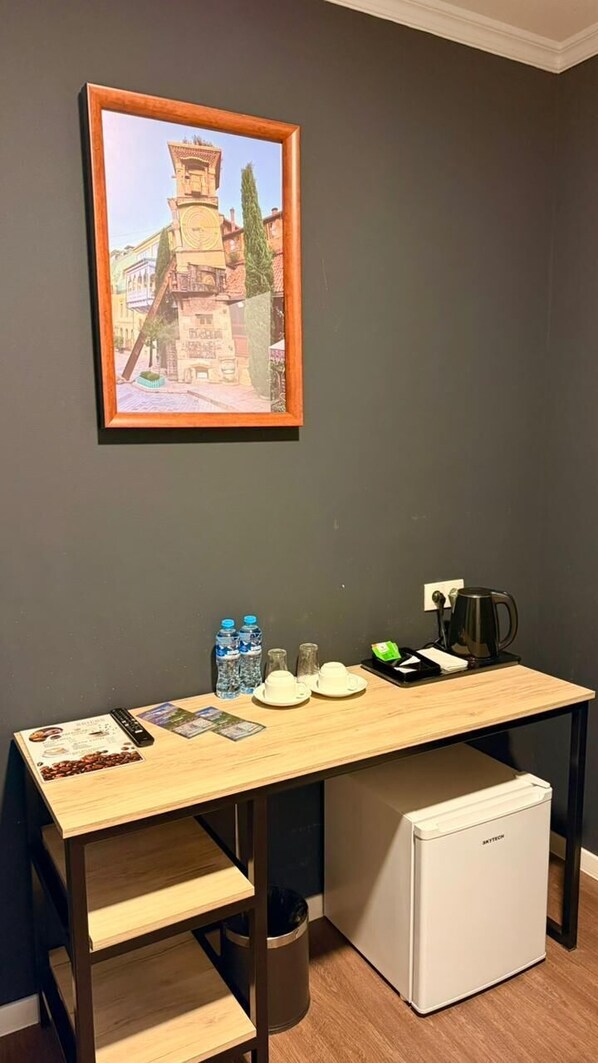 A compact room with a wooden desk, a small refrigerator, a framed picture on the wall, and a coffee maker.