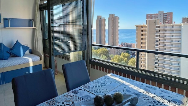 A balcony with a table set for dining, a sofa with cushions, and a view of tall buildings and the sea.