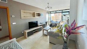 A modern living room with a flat-screen TV, a dining area with a table and chairs, and a balcony with a view of the city.