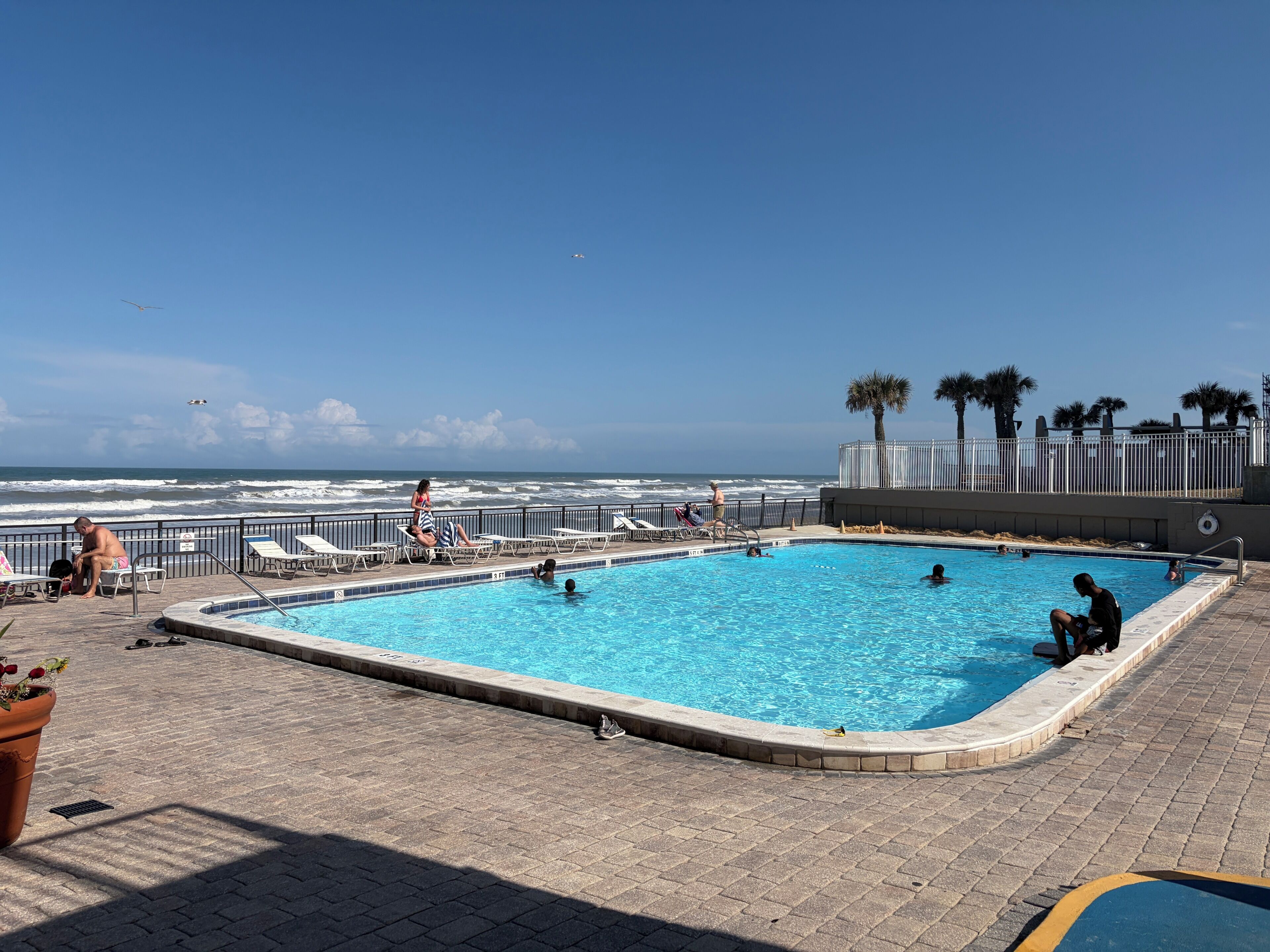 A pool area with a view of the ocean, lounge chairs, and a fenced deck.