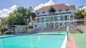 A large swimming pool with a clear blue water, surrounded by a stone wall and a covered area with a roof. - Malibu ocean View Studio (Montego Bay)