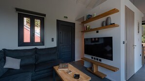 A modern living room with a dark-colored sofa, a wooden coffee table, a flat-screen TV mounted on a white wall, and a bookshelf with decorative items. - Agata - Two Bedroom Villa, Sleeps 4 (Fiskardo)