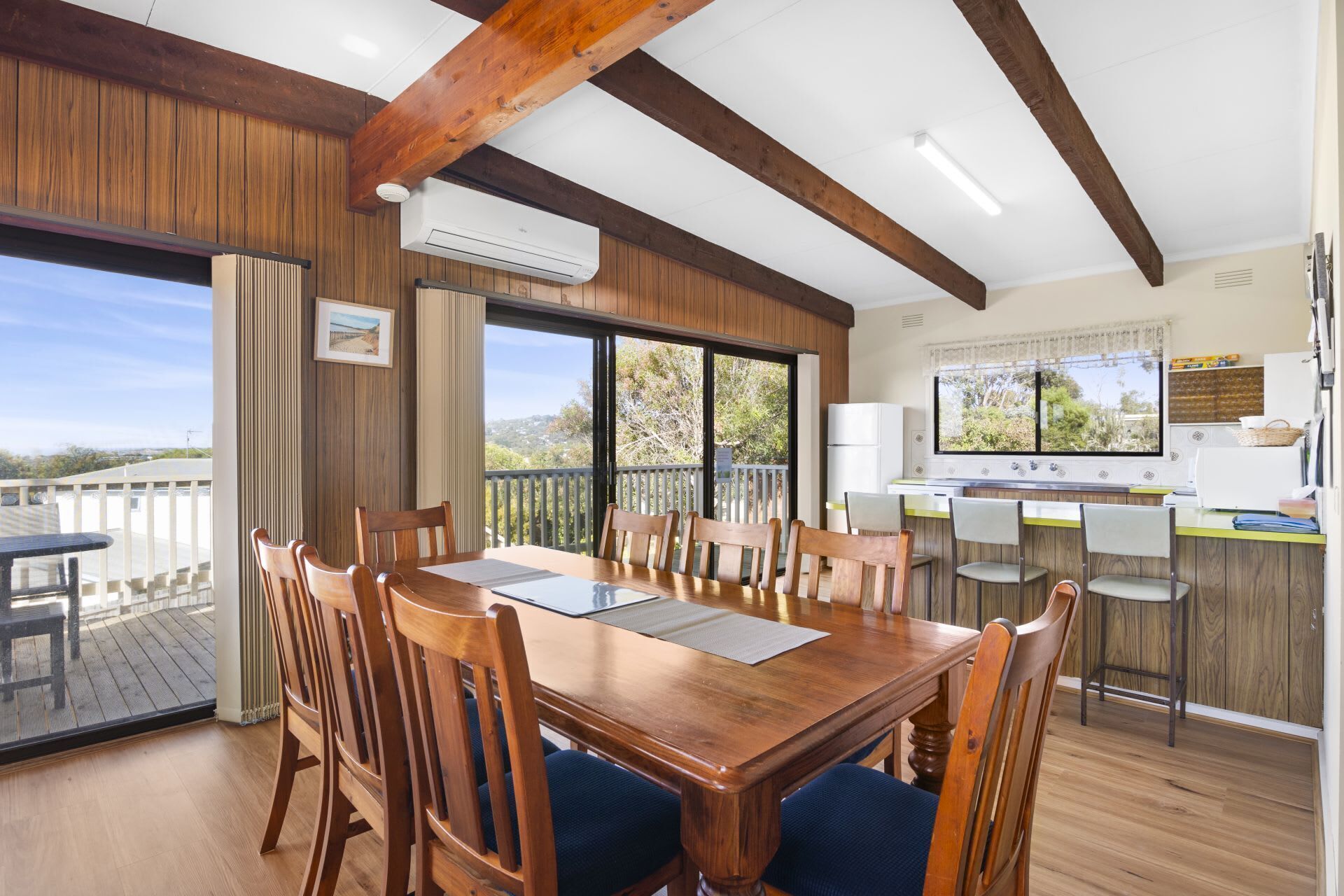 A dining area with a wooden table and chairs, a kitchen with a large window, and a view of the outdoors.