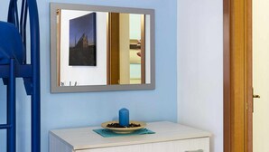 Quarto com parede azul clara, porta de madeira, cómoda branca com gavetas e espelho refletindo um edifício e uma porta.