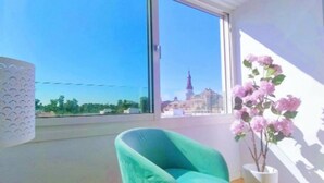 Ein Zimmer mit großem Fenster, einem Türkis-Armlehnstuhl und einem Blumentopf mit rosa Blüten.