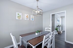 Un coin repas avec une table en bois, des chaises blanches, un luminaire suspendu et des œuvres d’art encadrées accrochées au mur.