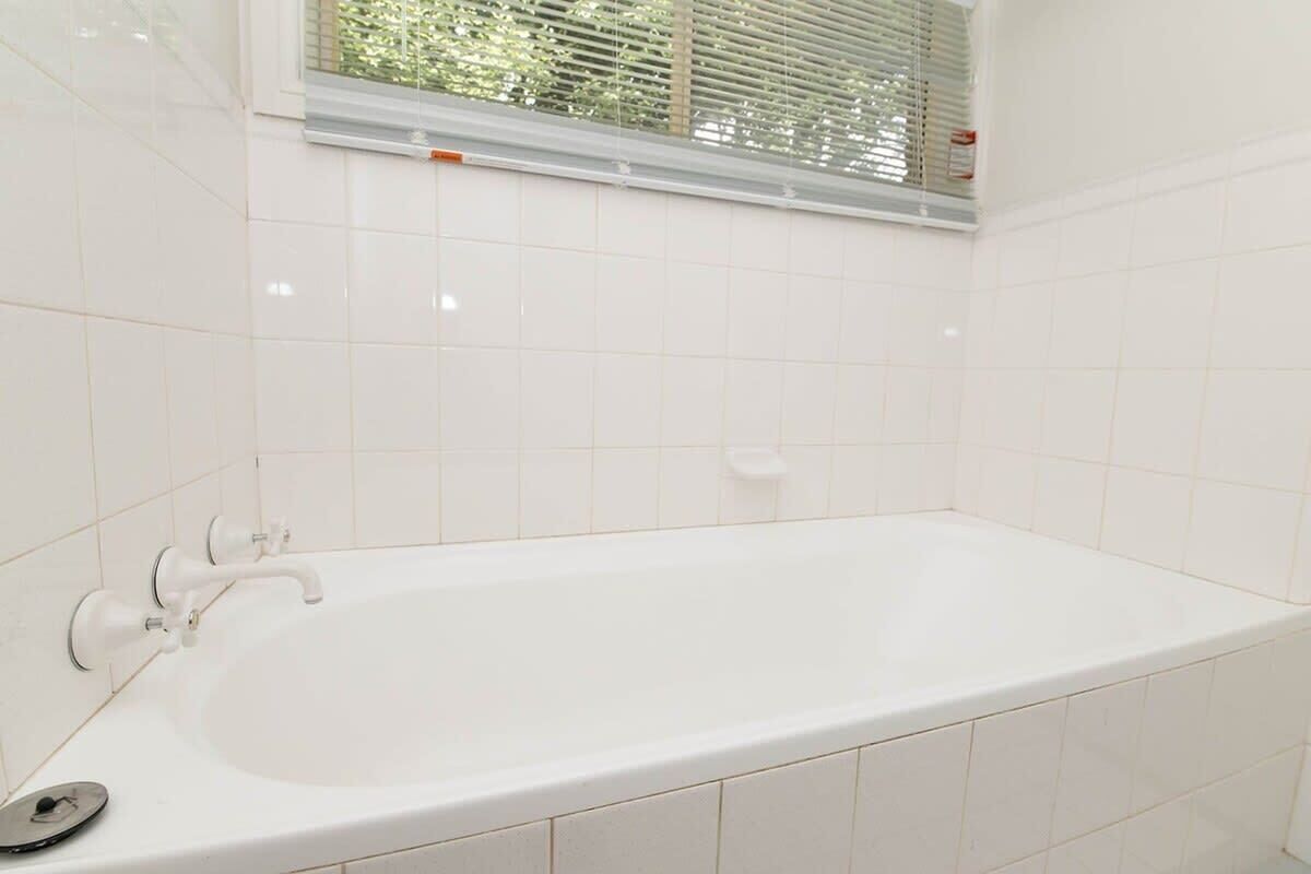 Een wit bad met een kraan en een zeepdispenser in een badkamer met witte betegelde wanden en een raam met zonwering.