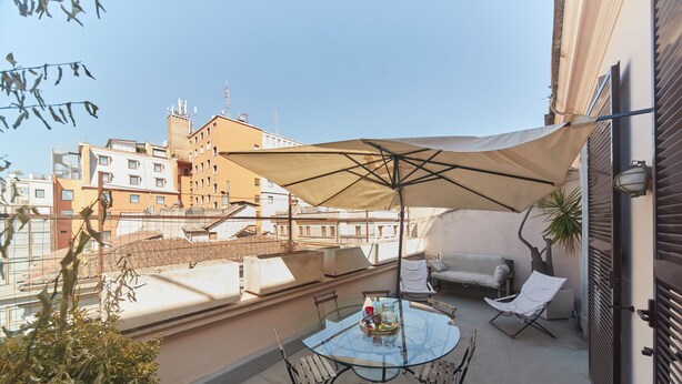 Une terrasse sur le toit avec une table en verre, des chaises, un grand parasol et un canapé.