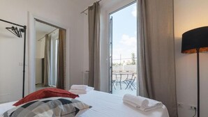 Une chambre d’hôtel avec un lit, un oreiller rouge, un oreiller à carreaux et une table avec des chaises devant la fenêtre.