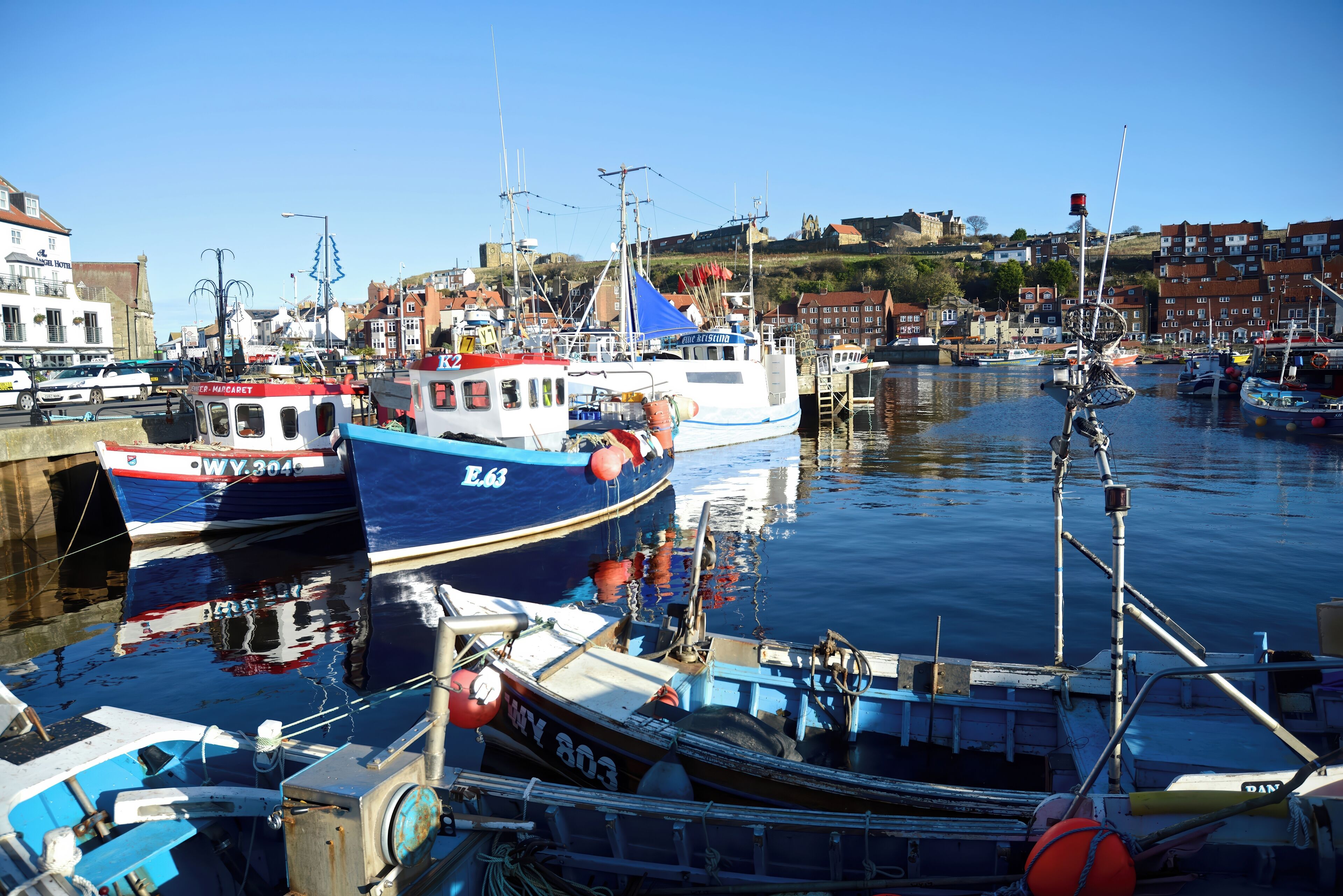 A marina with various boats docked, a clear sky, and buildings in the background.