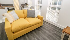 Un sofà groc amb coixins estampats, una taula de centre de fusta i una taula auxiliar de fusta en una habitació lluminosa amb finestres grans i una cuina al fons.