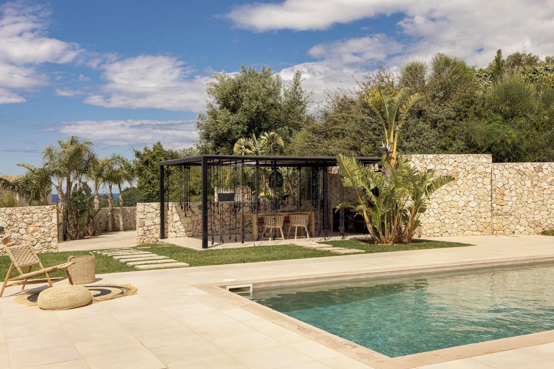 A pool area with a stone wall, a covered seating area, and tropical plants.