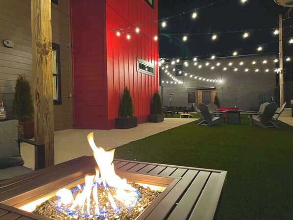 A backyard with a fire pit, outdoor seating, string lights, and a red building.