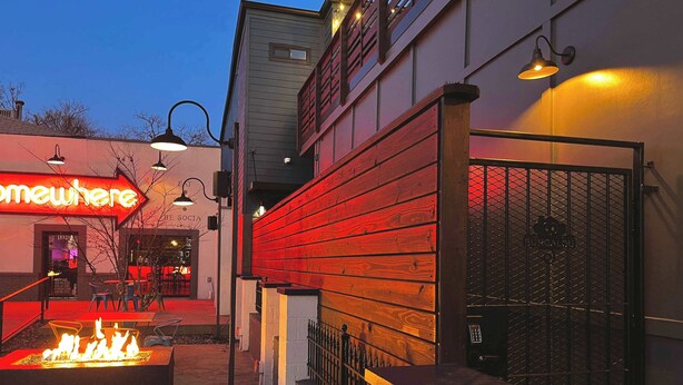 An outdoor seating area with a fire feature, a metal gate, and a building with a neon sign. - Swanky Suite @ Bungalou + Bourbon Bar + Gameyard (Louisville)