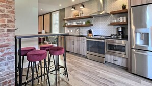 A modern kitchen with bar stools, stainless steel appliances, and a brick wall. - Swanky Suite @ Bungalou + Bourbon Bar + Gameyard (Louisville)