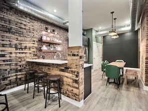 Une cuisine moderne avec un mur d’accent en briques, un îlot central doté de tabourets et un coin repas avec des chaises vertes.