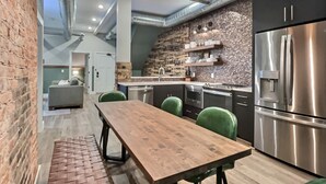 A modern kitchen with stainless steel appliances, a wooden dining table, green chairs, and a brick wall.