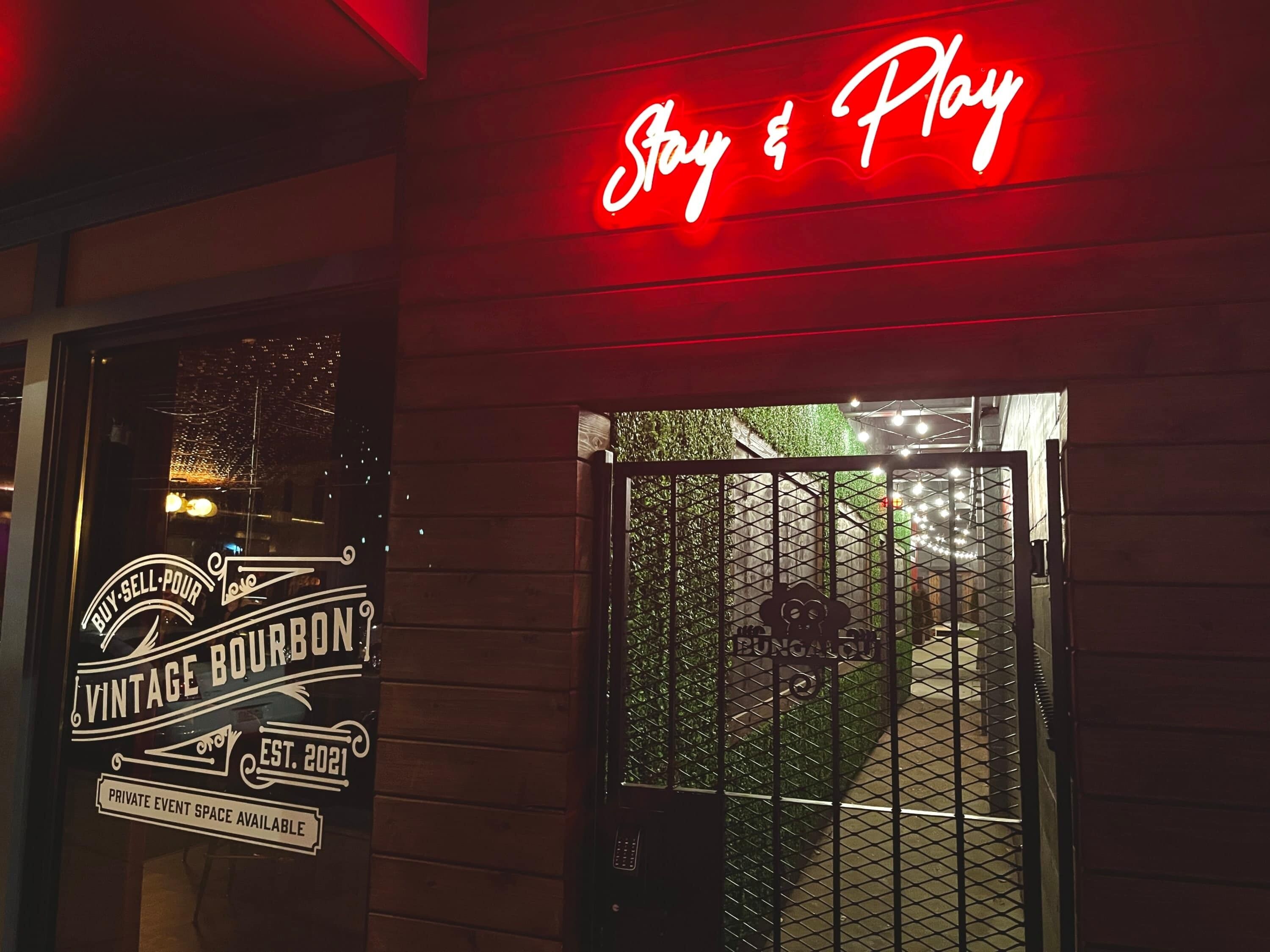 A storefront with a neon sign reading 'Stay & Play' and a window sign advertising 'Vintage Bourbon' with private event space.