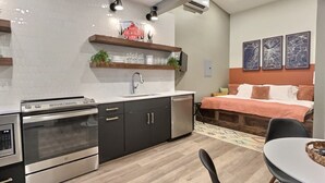 Una cocina compacta con electrodomésticos modernos, un comedor con mesa y sillas, y una cama con cabecera colorida.