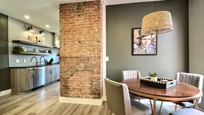 A modern kitchen with a brick accent wall, a dining area with a round table and chairs, and a hanging light fixture. - Swanky & Upbeat Suites @ Bungalou + Bourbon Bar (Louisville)
