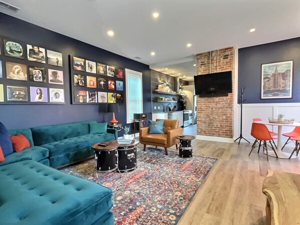 Una sala de estar con un sofá azul, un tapete estampado, un comedor con sillas rojas y una cocina al fondo.
