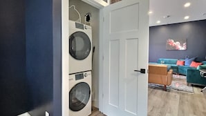 A laundry room with stacked washers and dryers, a white door, and a living area with a blue sofa and cushions. - Swanky & Upbeat Suites @ Bungalou + Bourbon Bar (Louisville)