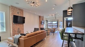 A modern living room with a brick wall, a flat-screen TV, a leather sofa, a dining area with stools, and a kitchen with a bar.