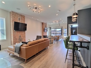 A modern living room with a brick wall, a flat-screen TV, a leather sofa, a dining area with stools, and a kitchen with a bar. - Rooftop Suite @ Bungalou + Bourbon Bar + Gameyard (Louisville)