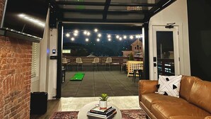A modern living room with a leather sofa, a coffee table with books and a plant, a TV mounted on a brick wall, and a view of an outdoor patio area with string lights and a green mat.