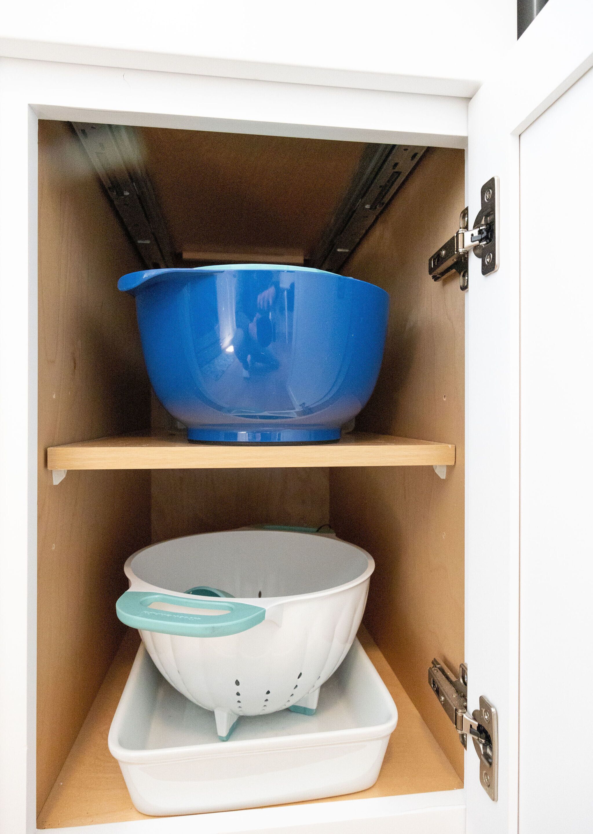 A kitchen cabinet with two shelves, one holding a blue bowl and the other a white bowl with a teal lid.