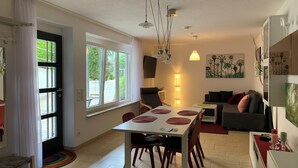 A modern living room with a dining area, a sofa, a coffee table, and a TV. There is a window with curtains, a lamp, and a painting on the wall.