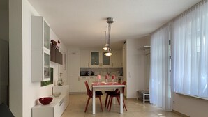 A modern living room with a sofa, a dining table with chairs, a kitchen in the background, and a large window with curtains.