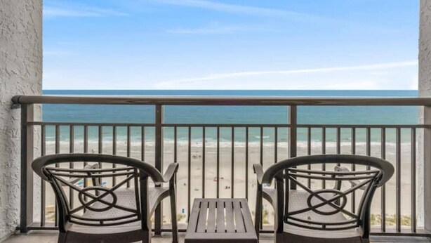Ein Balkon mit zwei Stühlen und einem kleinen Tisch, der Blick auf einen Strand und das klare Meer gewährt.