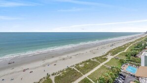 Ein Strand mit freiem Blick auf den Ozean und einem Poolbereich mit Liegestühlen und Sonnenschirmen.