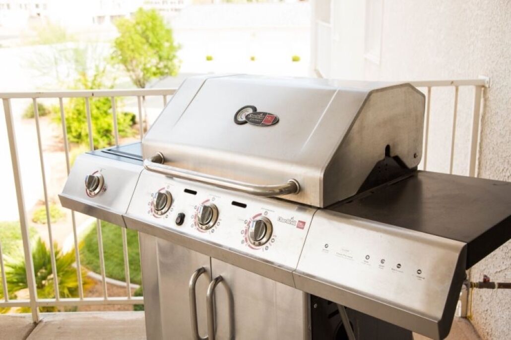 A stainless steel barbecue grill with four burners and a digital control panel, located on a balcony with a railing.