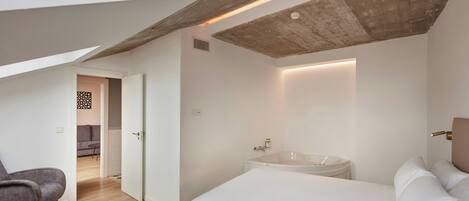 A modern bedroom with a large bed, a sitting area with a chair, and a bathroom with a bathtub.