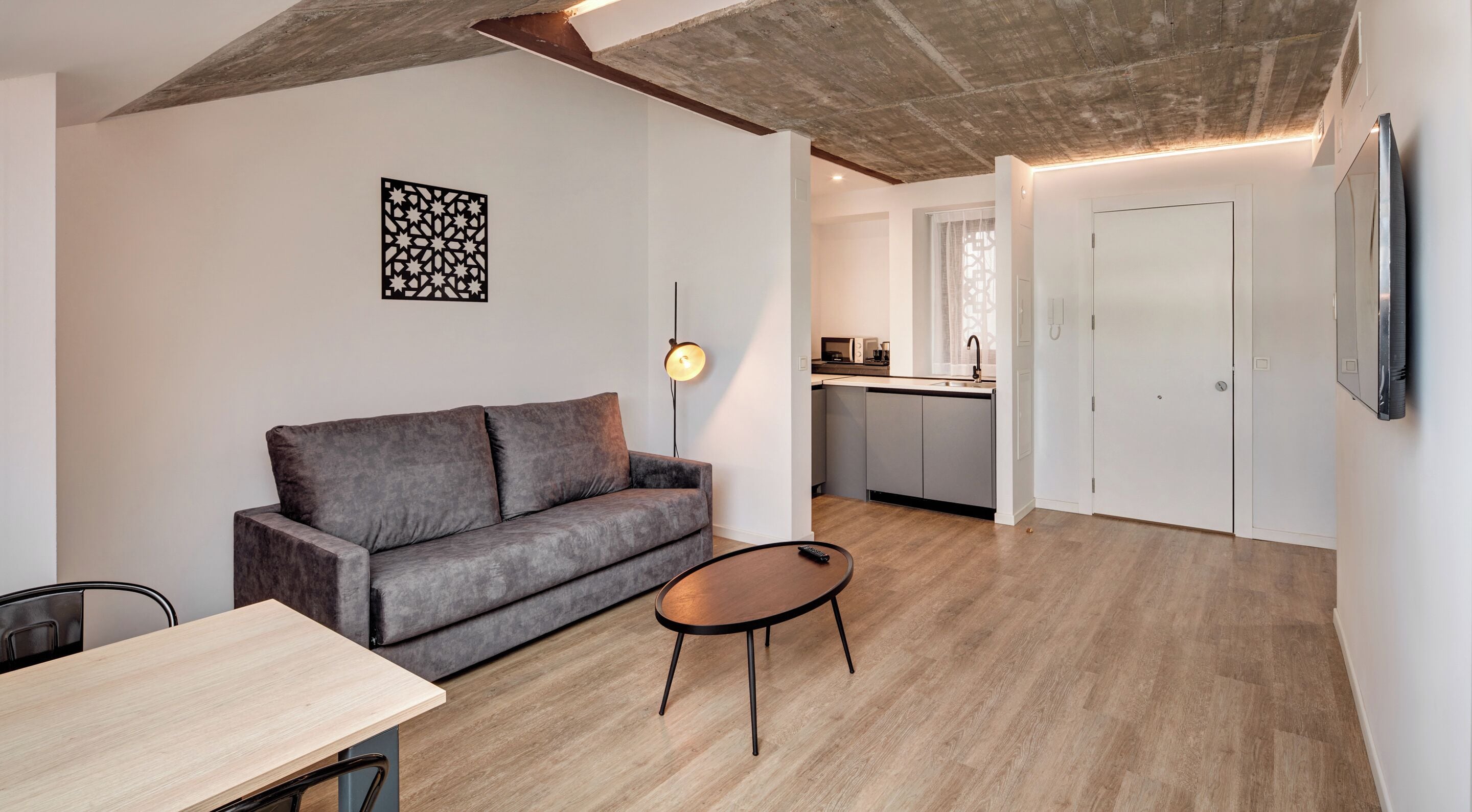 Un salon moderne avec un canapé gris, une petite table ronde et une cuisine en arrière-plan.