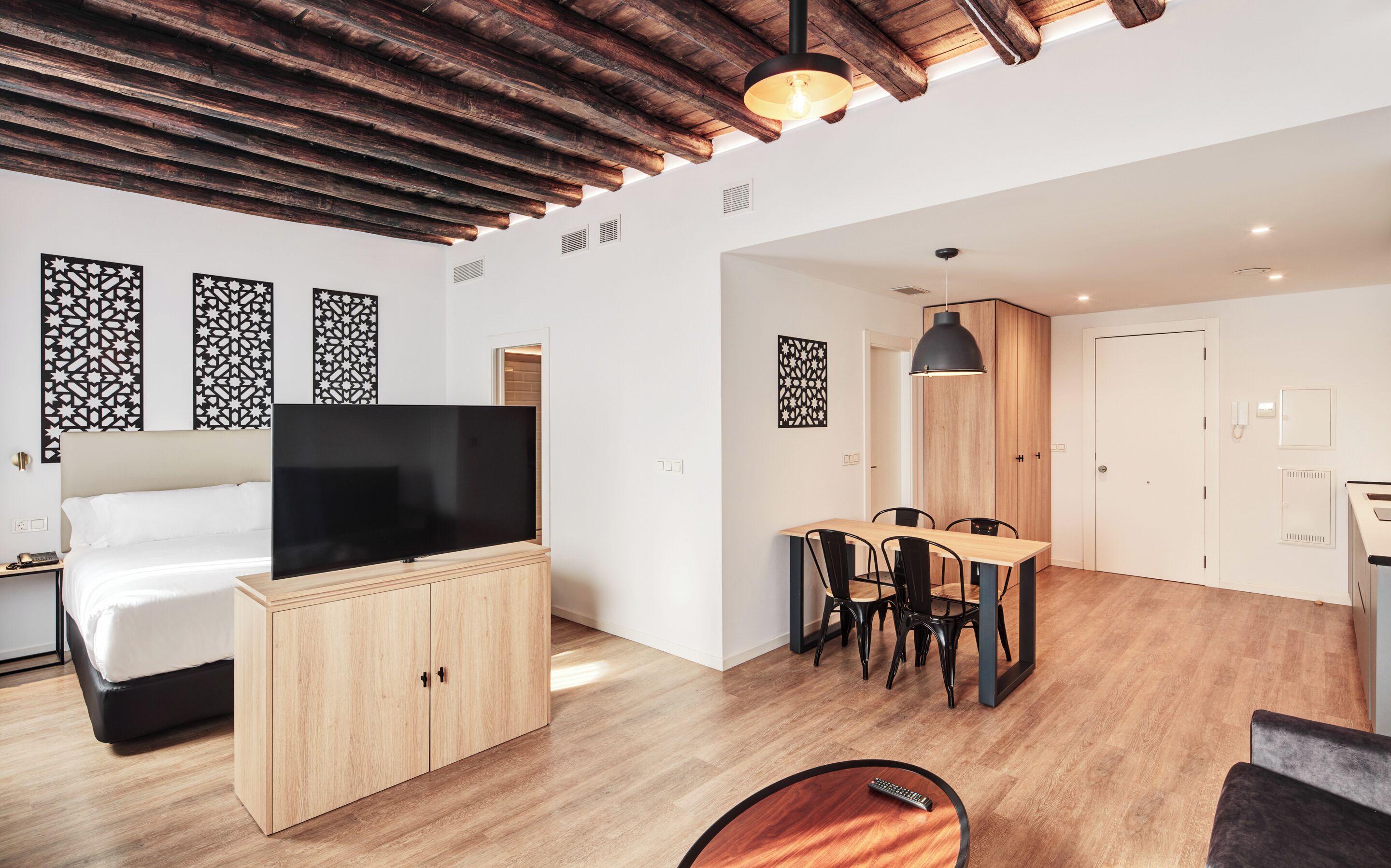 Ett modernt hotellrum med ett träpanelat tak, en platt-TV, en säng, en matplats med bord och stolar samt dekorativa väggpaneler.