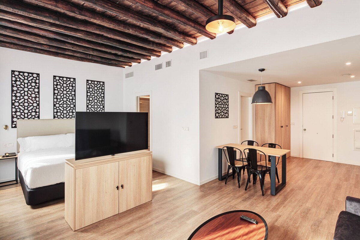 Ein modernes Hotelzimmer mit Holzboden, einem Flachbildfernseher, einem Bett, einem Essbereich mit Tisch und Stühlen sowie dekorativen Wandverkleidungen.
